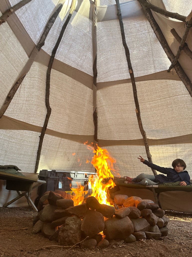 Inside the teepee: a campfire in a ring of river rocks, canvas walls, and Bastion on a green camping cot giving a peace sign.