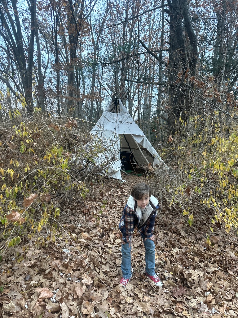 A canvas teepee in a leaf-strewn clearing among bare trees, with Bastion in a plaid shirt and red sneakers in the foreground.